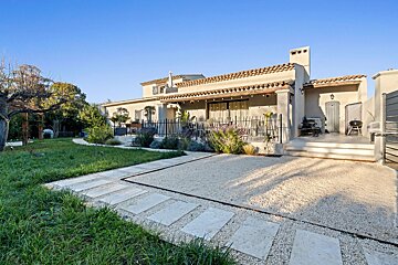 Sunny exterior of a Mediterranean-style house with a garden, gravel path, and patio. Tiled roof and stucco walls stand out against a blue sky.