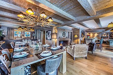 Luxurious rustic wooden chalet interior with a grand dining table, antler chandeliers, cozy fireplace, and lounge area, exuding warmth.