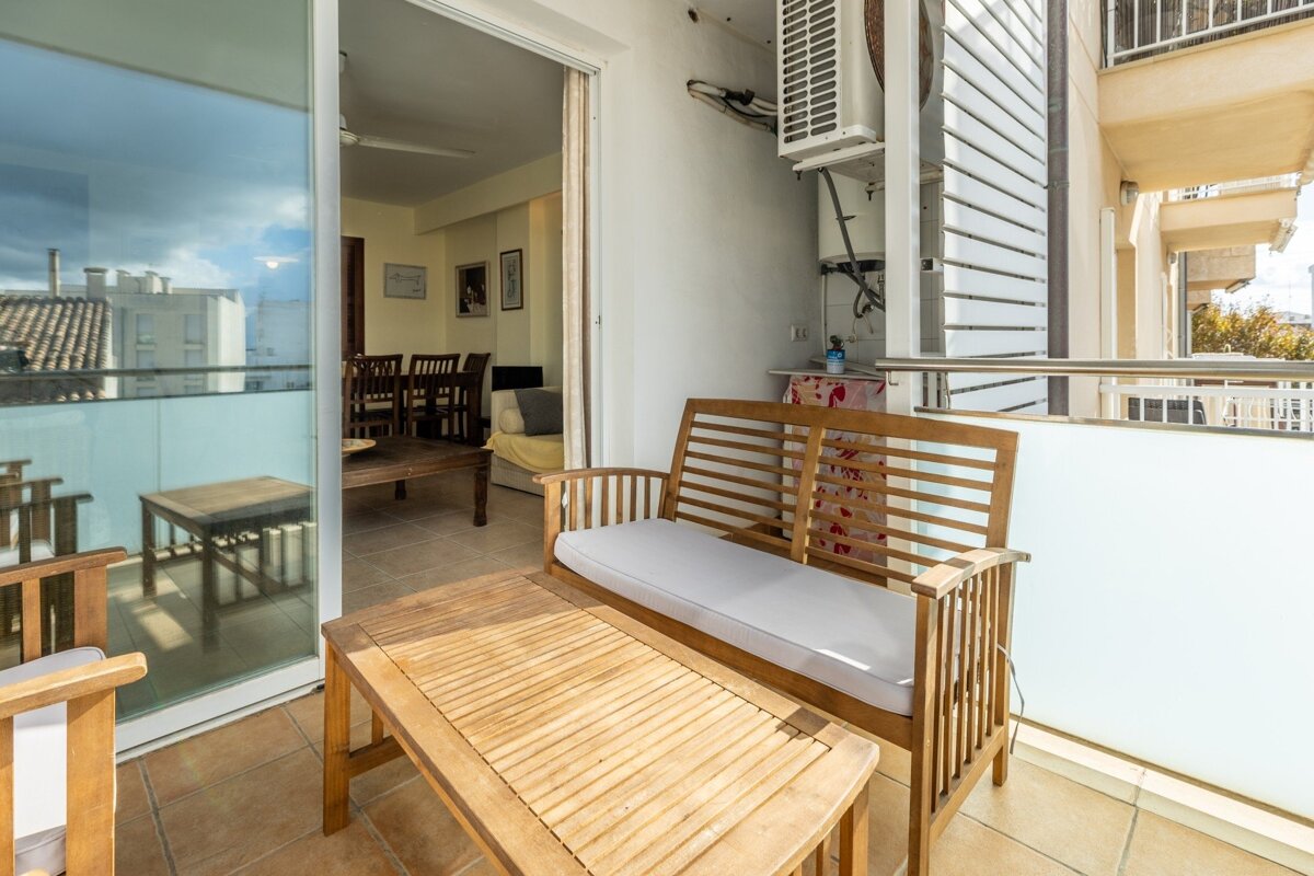 A sunlit balcony with wooden outdoor furniture, an AC unit on the wall, and a view into a living room with a dining table, plus distant buildings.