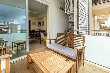 A sunlit balcony with wooden outdoor furniture, an AC unit on the wall, and a view into a living room with a dining table, plus distant buildings.