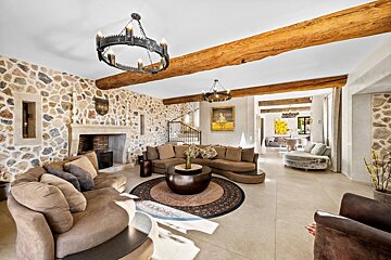 A grand living room featuring stone walls, rustic wooden beams, and two medieval-style chandeliers above curved brown sofas and a fireplace.