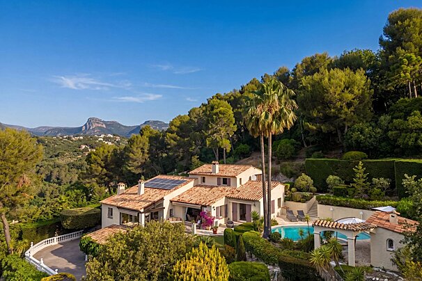 A sprawling house with solar panels, a pool, and lush gardens, set against a backdrop of mountains under a clear blue sky.