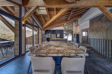Spacious chalet interior with exposed wood beams, a large granite dining table, modern kitchen, and stunning mountain views through expansive windows.