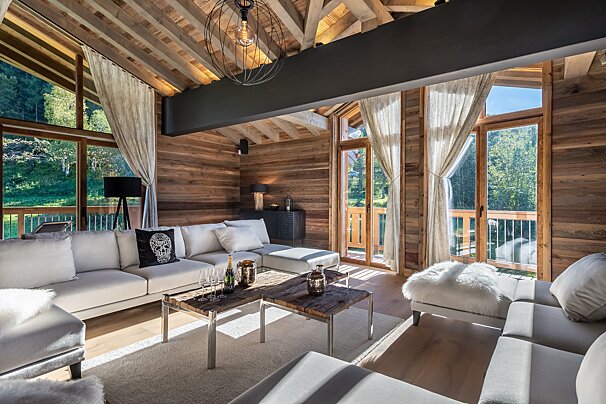 Cozy, modern chalet living room with wooden walls, white sofas, rustic coffee tables, and large windows showcasing a scenic outdoor view.