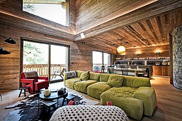 Modern rustic chalet interior with wood walls/ceiling, a large green sofa, red armchair, open kitchen, and balcony views.