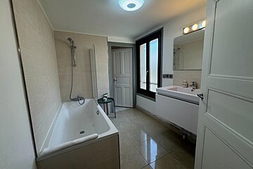 A bright, neutral-toned bathroom with a tub/shower combo, vanity sink, mirror, and window. Tiled floor/walls. Door opens to another room.