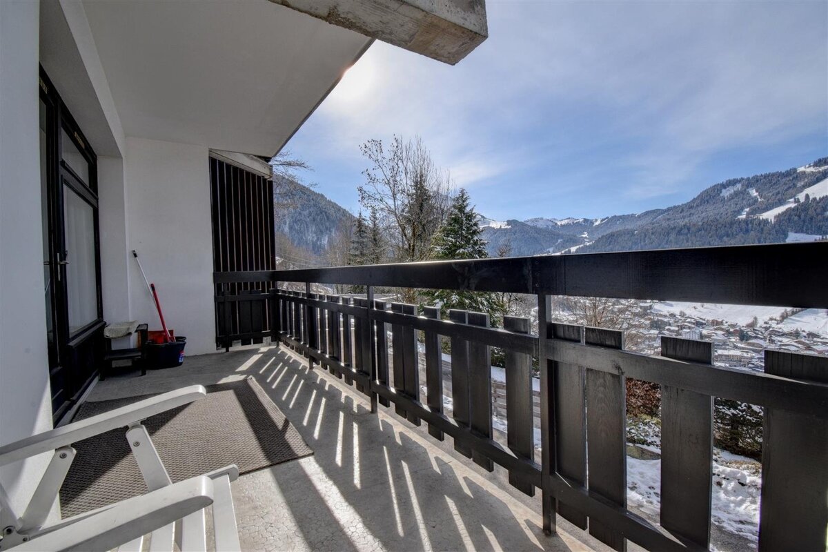 Sunny balcony with a dark wooden railing overlooking a beautiful snow-covered mountain valley.