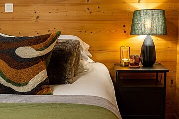 Cozy bedroom with a wood-paneled wall, textured pillows on the bed, and a warm lamp lit on a black nightstand.