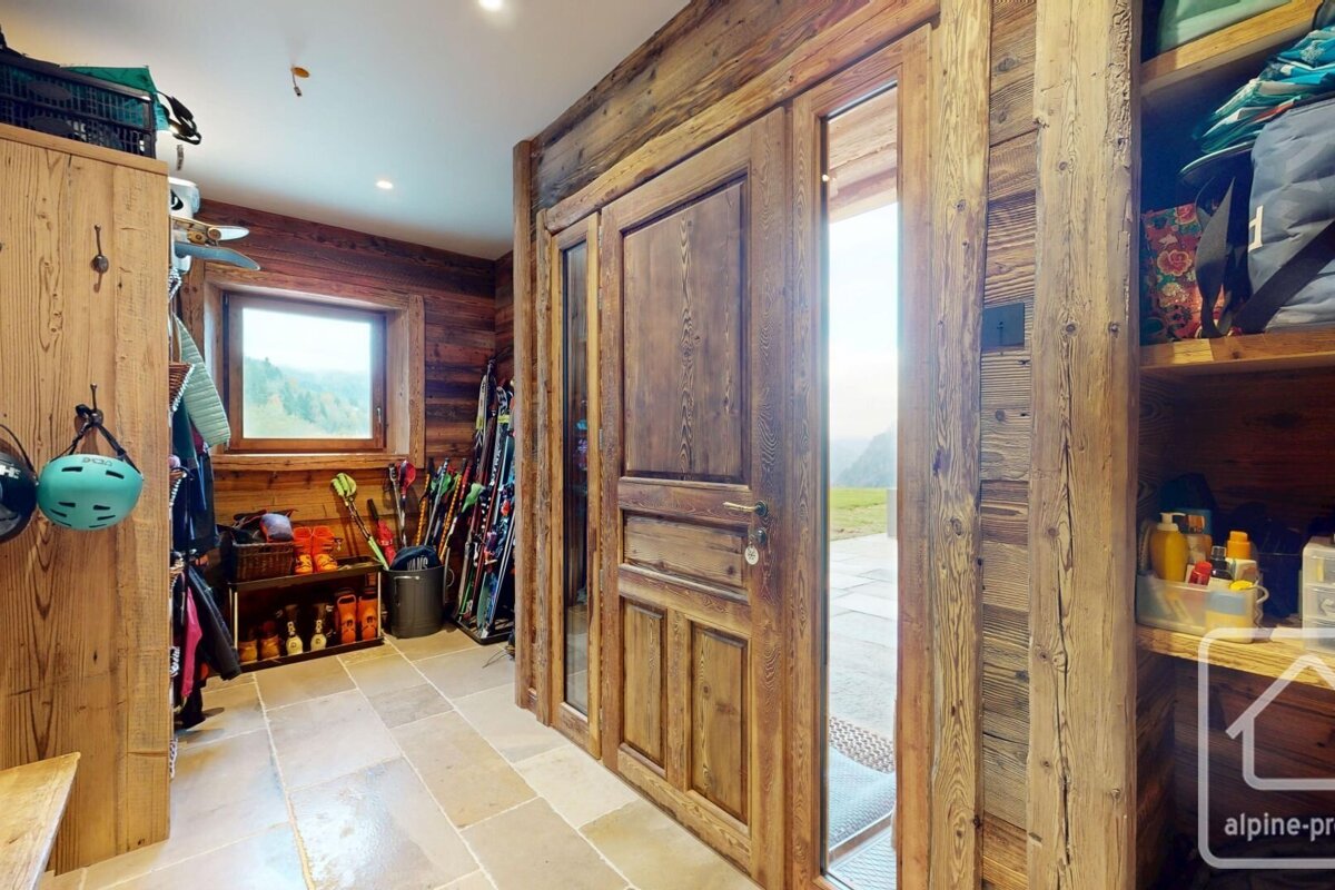 A rustic wooden ski lodge entryway with stone floor, packed with ski gear. A large wooden door and window offer mountain views.