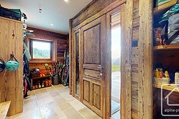 A rustic wooden ski lodge entryway with stone floor, packed with ski gear. A large wooden door and window offer mountain views.