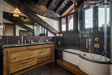 Rustic modern bathroom with dark exposed wooden beams, a wooden double vanity, and a sleek glass-enclosed shower/tub. Dark tiles complete the design.