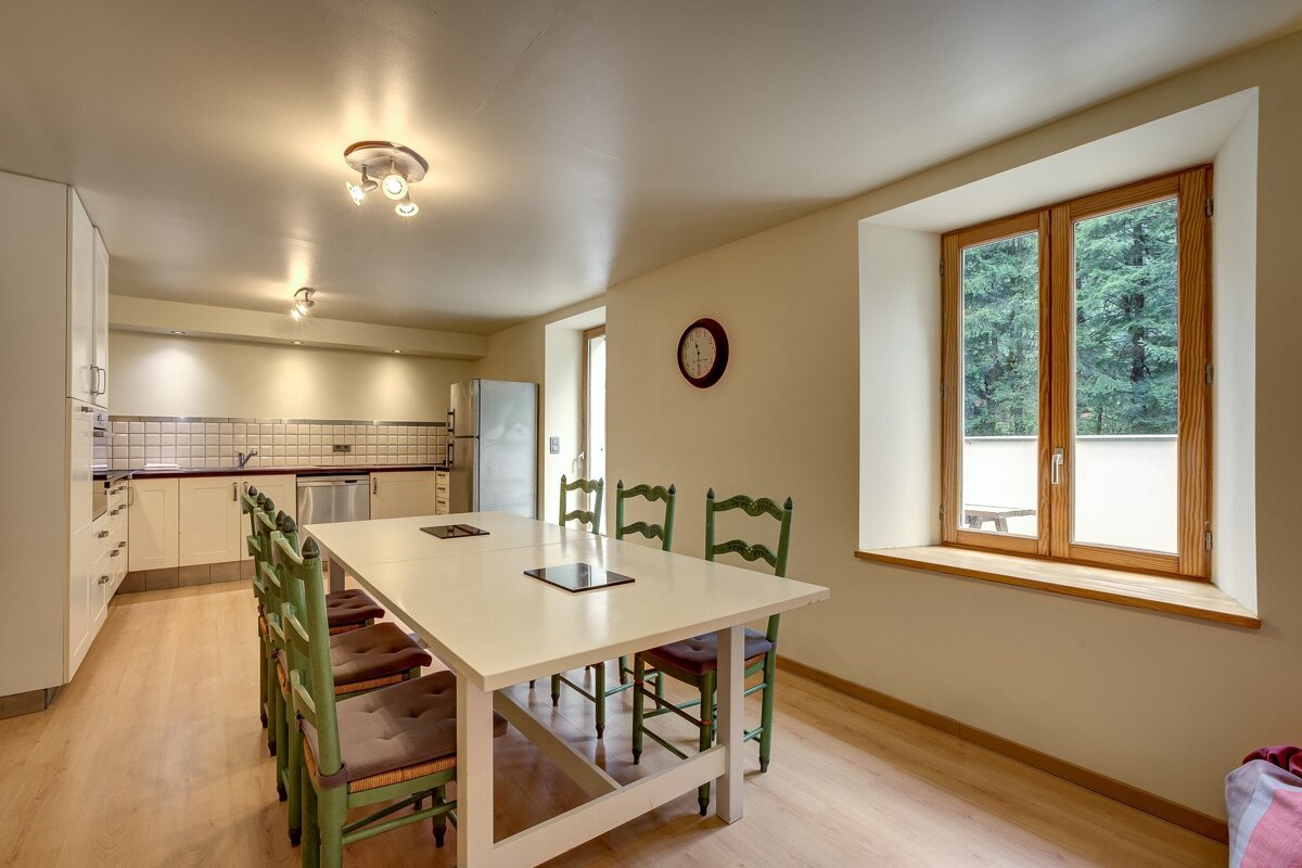 A bright kitchen-diner featuring a long white table, green chairs, modern appliances, and a window with a forest view.