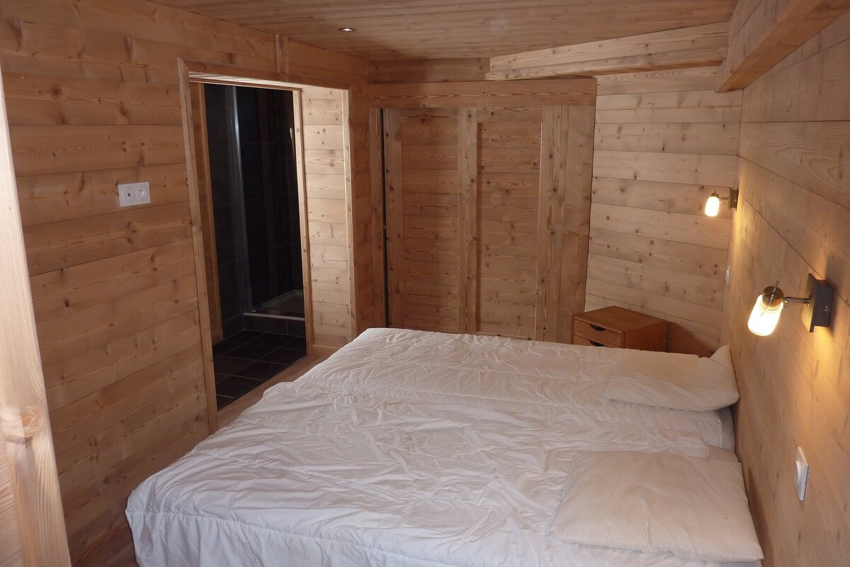A wood-paneled cabin bedroom with a large bed in white sheets, glowing wall lamps, and a doorway to a bathroom.