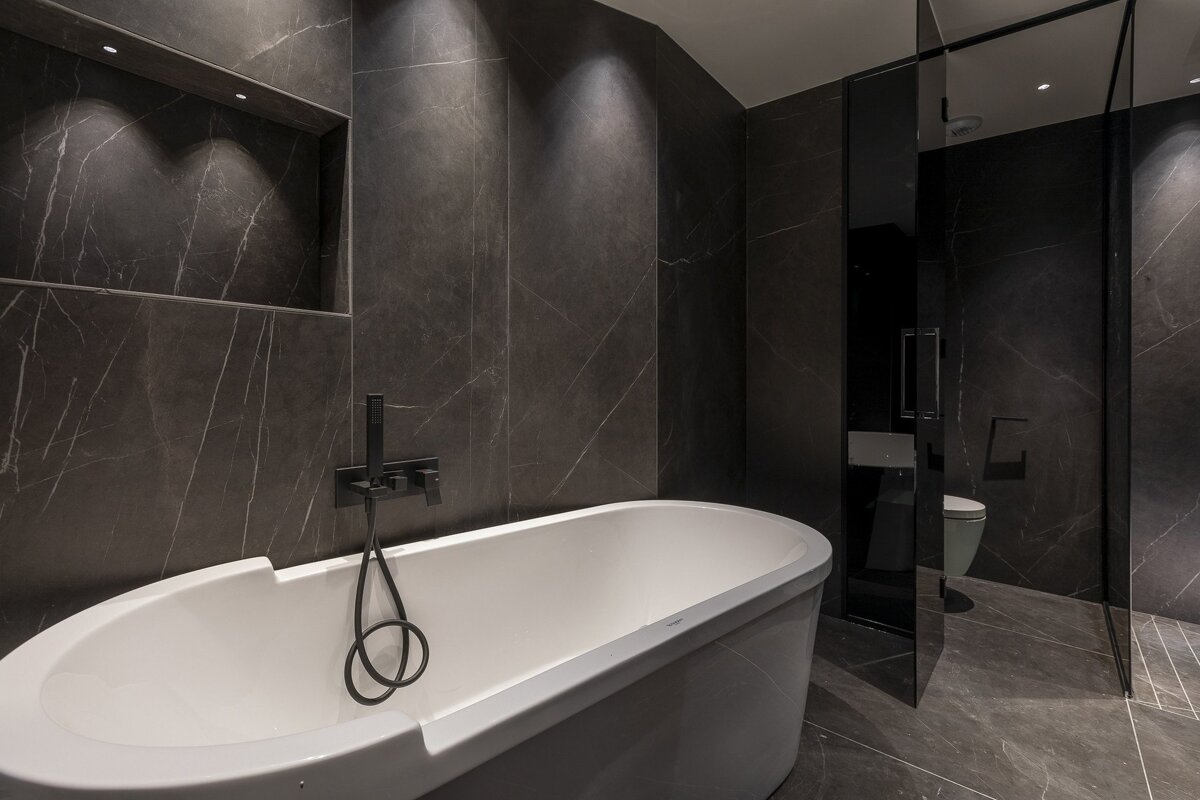 Modern dark-themed bathroom with a white freestanding tub, black fixtures, and dramatic dark marble tiles.