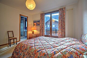 A cozy bedroom with a colorful patterned bedspread, glass doors leading to a balcony with a snowy mountain view, and an en-suite bathroom.