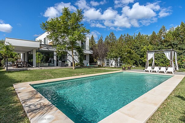 A modern white house with a large swimming pool and green lawn. Tall trees surround the property, and a poolside gazebo offers shade.