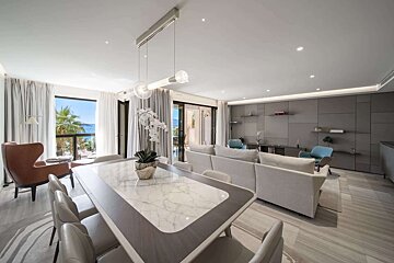 A modern open-plan living/dining room with ocean views through large windows, featuring a marble-top table, neutral sofas, and a gray accent wall.