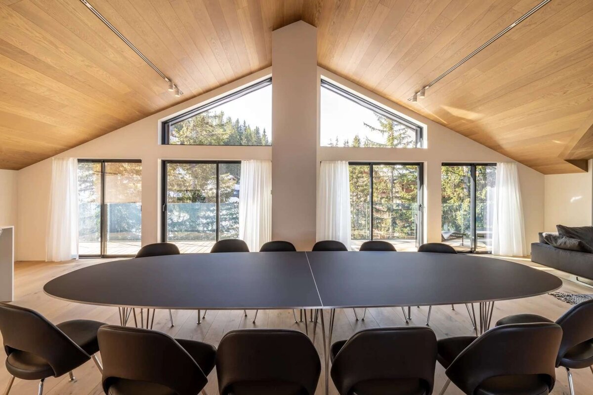 Bright, modern room with a large oval dining table & chairs, vaulted wooden ceiling, and panoramic windows overlooking a lush forest.