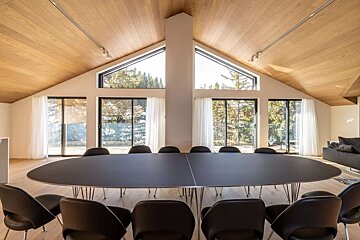 Bright, modern room with a large oval dining table & chairs, vaulted wooden ceiling, and panoramic windows overlooking a lush forest.