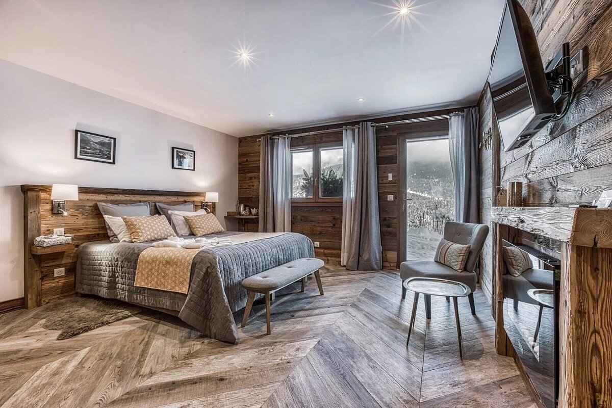 A cozy, rustic chalet bedroom featuring a large bed, wooden decor, and herringbone floor, with a stunning winter mountain view from the window.