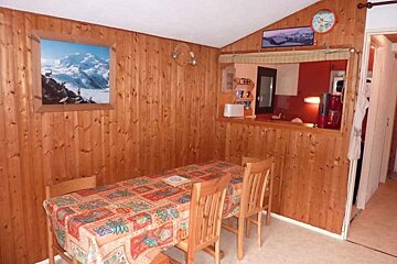 A rustic, wood-paneled room with a dining table and four chairs, facing a kitchenette through an open counter. Mountain artwork adorns the walls.