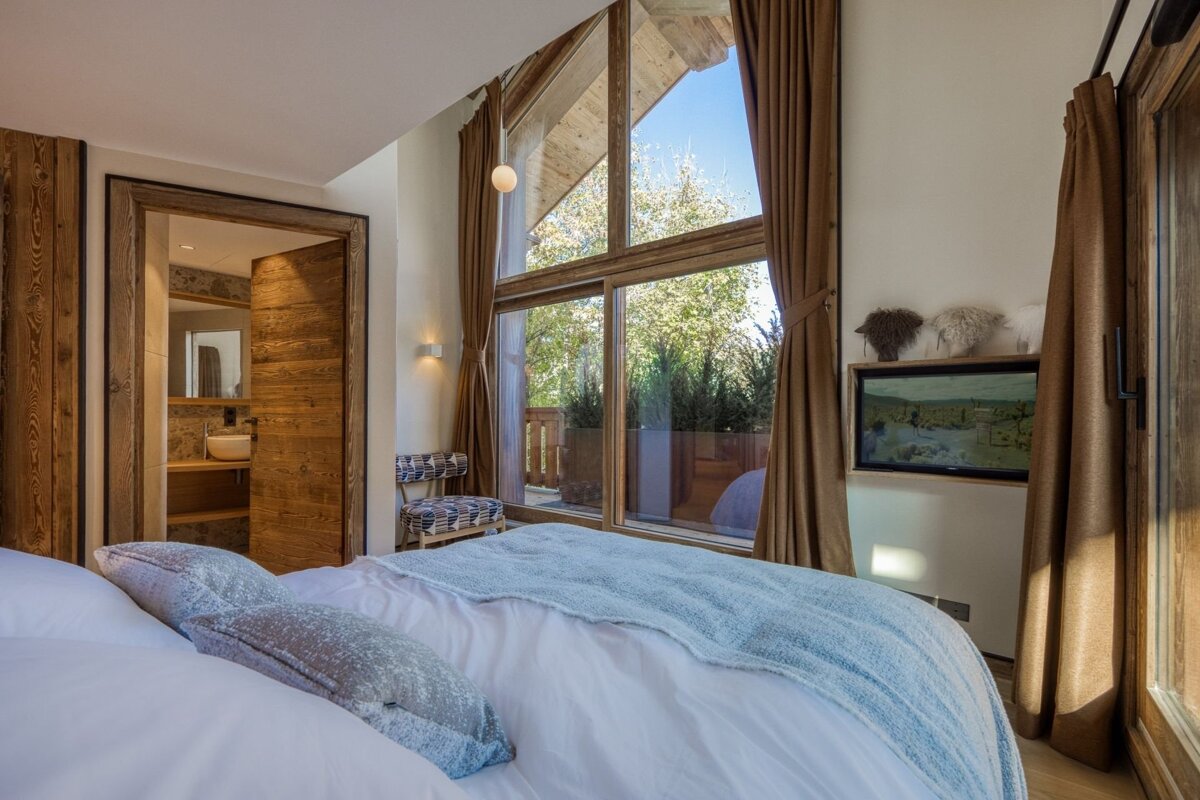 A warm, rustic bedroom with a large window offering nature views, a cozy bed, and an open door to a modern bathroom.