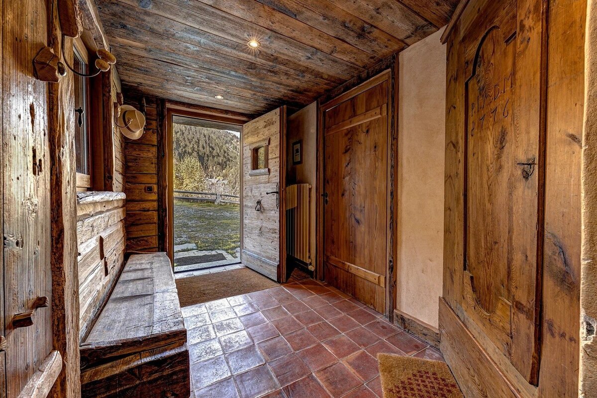 A cozy, rustic wooden entryway with a terracotta tiled floor. The open door offers a beautiful view of a forested mountain landscape.