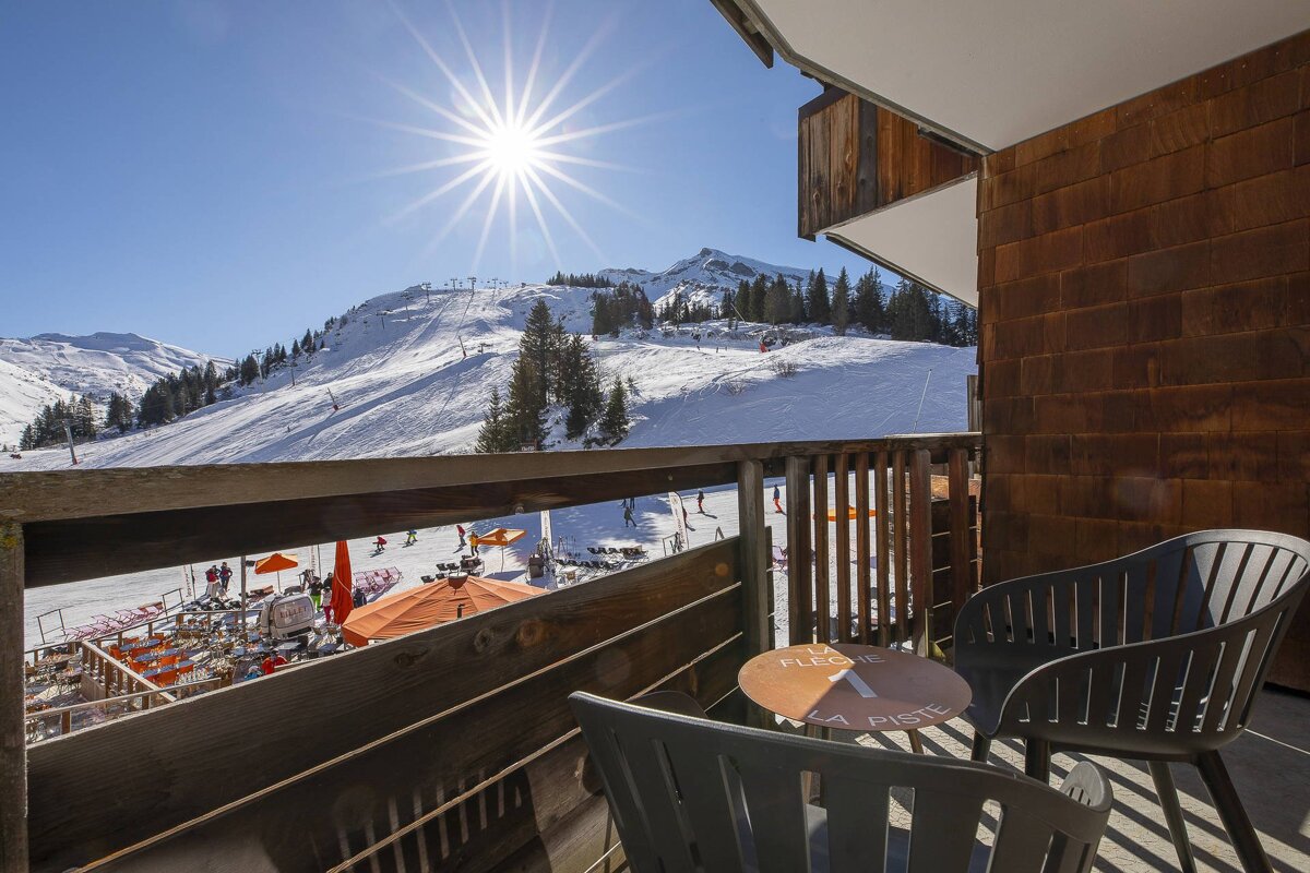 Sunny balcony view of a bustling ski resort. Snow-covered mountains, skiers, an outdoor cafe, and a table with chairs are visible.