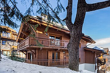 A rustic wooden chalet with balconies, surrounded by snow and a large tree, set against a bright blue sky in a winter mountain scene.