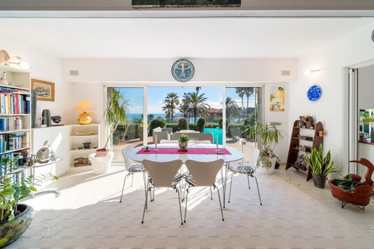 A bright, modern dining room with a white table, chairs, and large glass doors opening to a sunny outdoor view of palm trees.