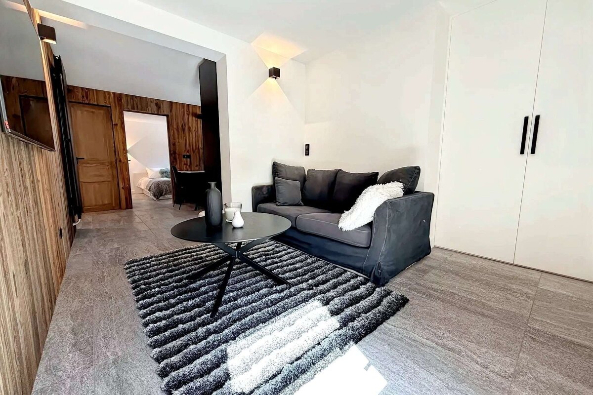 Cozy modern living room with a dark grey sofa, black coffee table, and striped rug. White walls, wood accents, and a view into a bedroom.