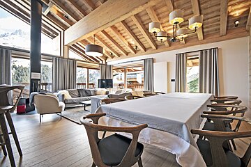 Spacious, modern chalet with open-plan living/dining, exposed wooden beams, and panoramic mountain views through large windows.