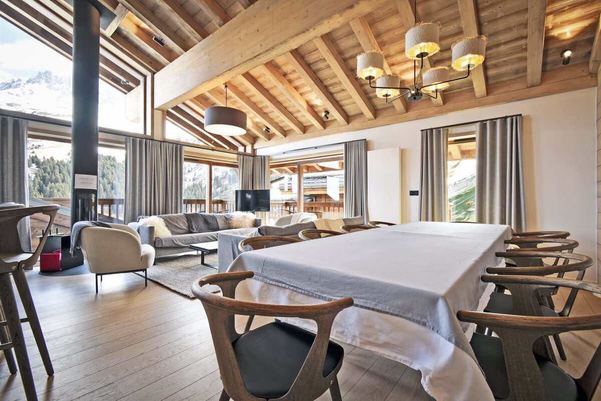 A rustic modern alpine chalet with exposed wooden beams, a large dining table, a cozy living area, and snowy mountain views.