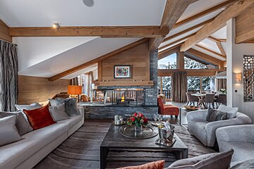 Luxurious alpine chalet living room with high wooden beams, a stone fireplace, plush sofas, and snowy views outside.