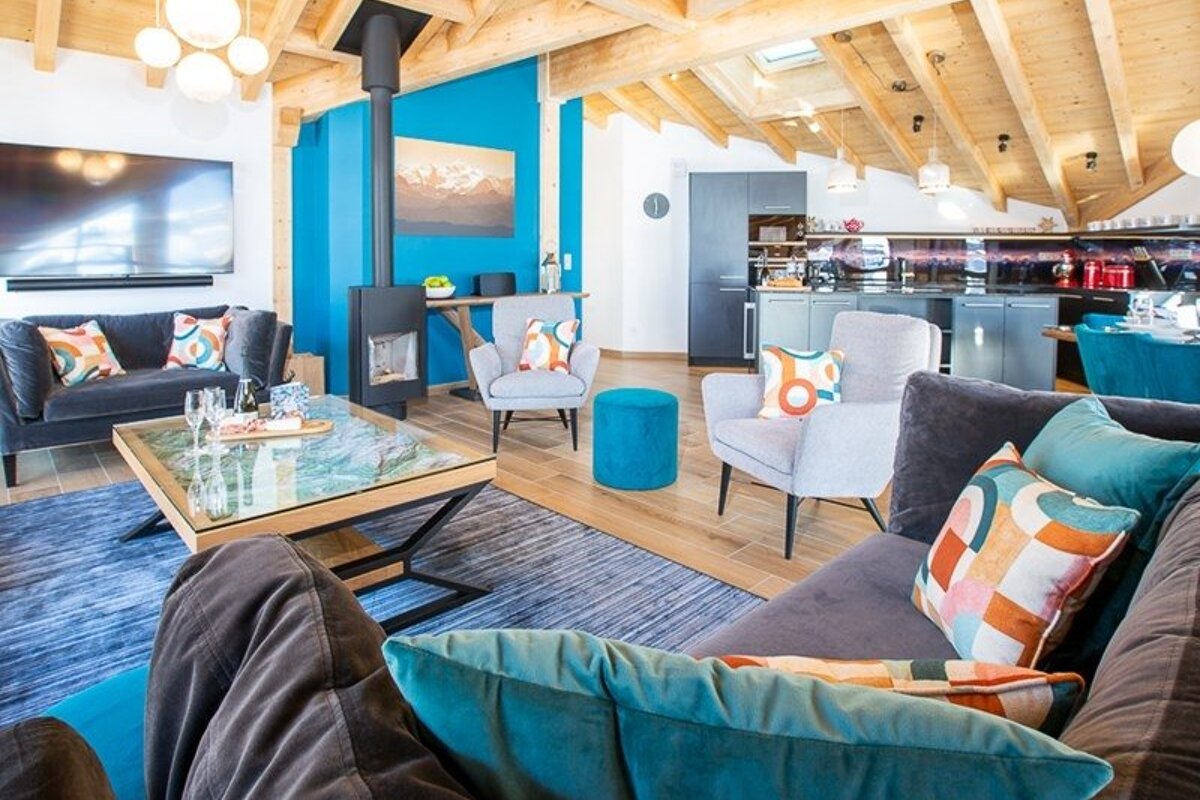 Cozy modern chalet living room with exposed wooden beams, a blue accent wall, colorful pillows, and an open kitchen.