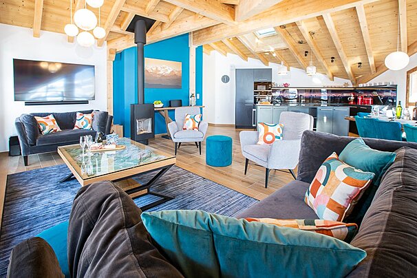 Stylish open-plan chalet living room with a bright blue wall, wooden beams, fireplace, and integrated kitchen area.