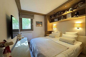 Cozy bedroom with a white bed, wooden decor, a TV, a window, and shelves full of plush toys above the headboard, lit by warm sconces.