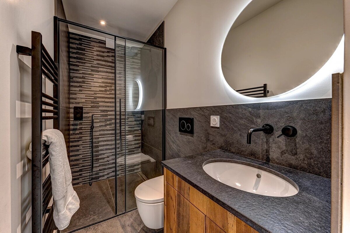 A sleek, modern bathroom featuring a glass shower, toilet, stone countertop with a sink, and a backlit oval mirror. Dark tones and wood accents create a stylish space.