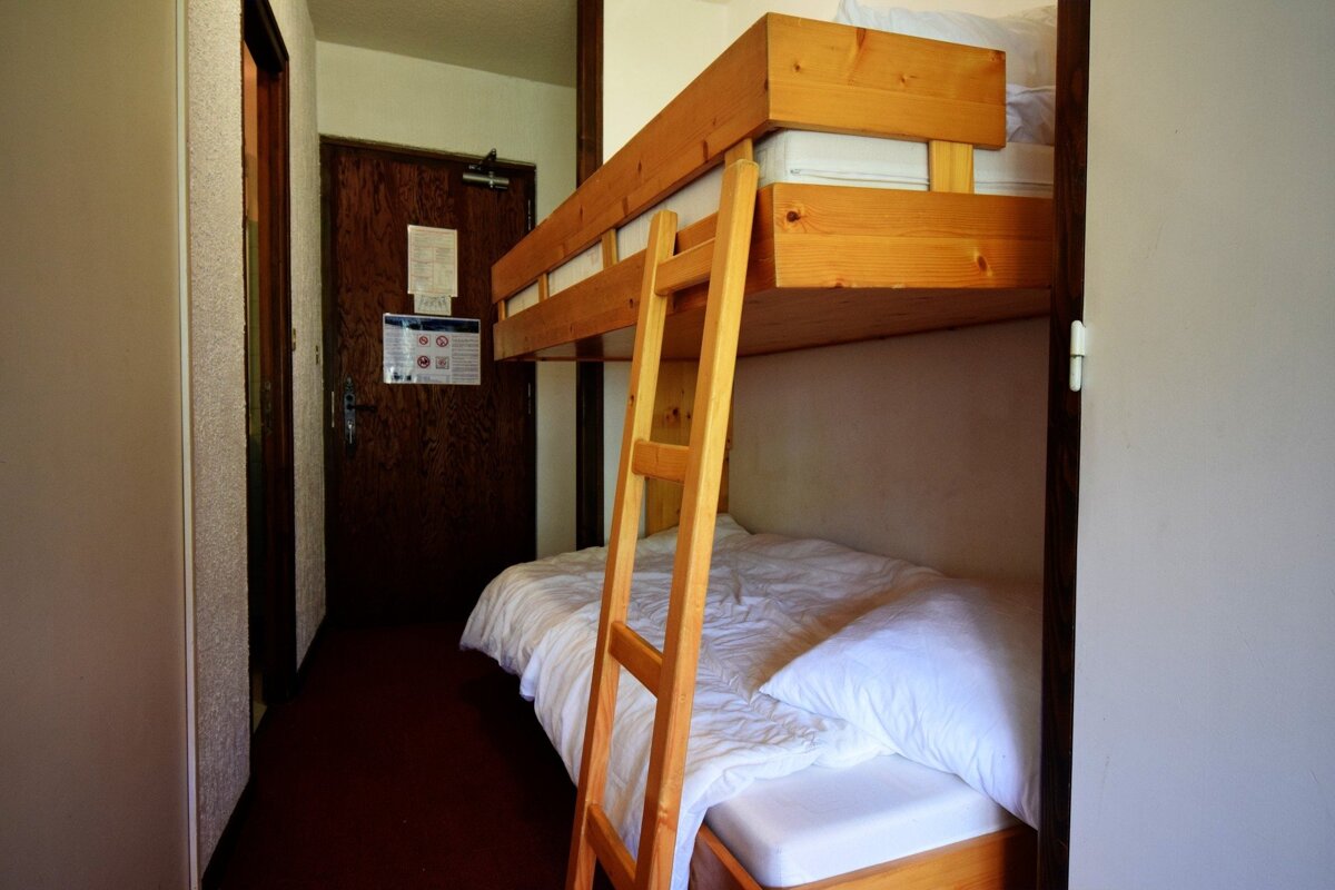 A narrow room with a wooden bunk bed, white bedding, and a ladder, next to a brown wooden door.
