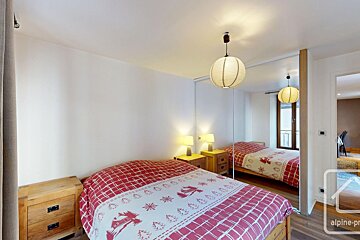 Cozy bedroom with red/white patterned bedding, wooden nightstands, lamps, and a mirrored wall reflecting the room, a window, and another room.