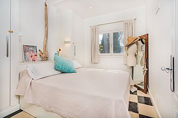 A bright, cozy bedroom featuring a white double bed with a teal pillow, a window with curtains, and a dress form with a hat beside a mirror. The room has built-in white cabinets and a black and white