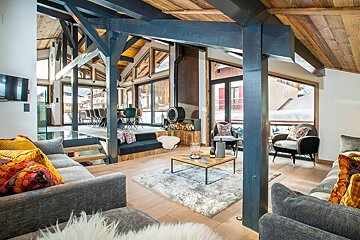 Cozy chalet living room with blue wooden beams, grey sofas, a fluffy rug, and large windows overlooking a snowy landscape.