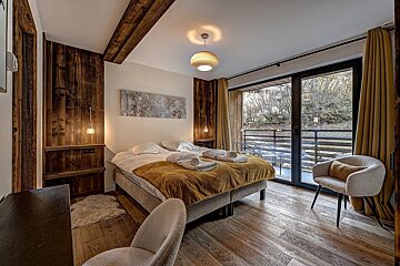 A cozy, modern rustic bedroom with a large bed, wooden walls, floor, and ceiling beams. Features a balcony overlooking nature.