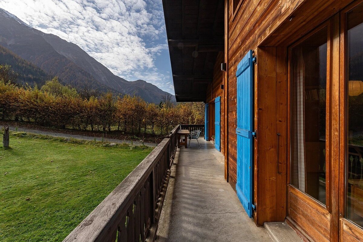 A wooden chalet balcony with vibrant blue shutters overlooks a green lawn, autumn trees, and majestic mountains under a cloudy sky.