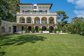 Luxurious Mediterranean villa featuring arched balconies, stone walls, and a sprawling green lawn under a bright blue sky.