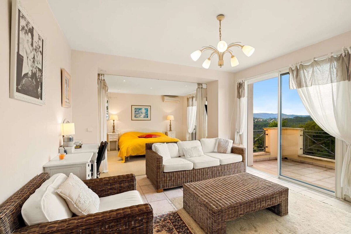 A bright, open-concept room features a living area with wicker furniture, a separate bedroom with a yellow bed, and a balcony with a scenic mountain view.