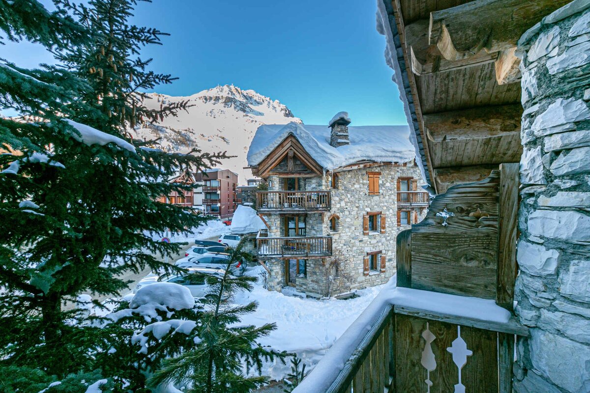 Charming stone chalet in a snowy mountain village, seen from a balcony with snow-dusted pines and a majestic peak under a clear blue sky.