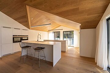A modern kitchen with a striking slanted wooden ceiling, white minimalist cabinets, two islands, and large sliding glass doors. Wooden floors complete the spacious, bright design.