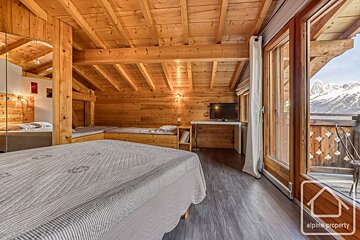 A rustic, all-wood loft bedroom features beds, a TV, and glass doors opening to a snowy mountain view.
