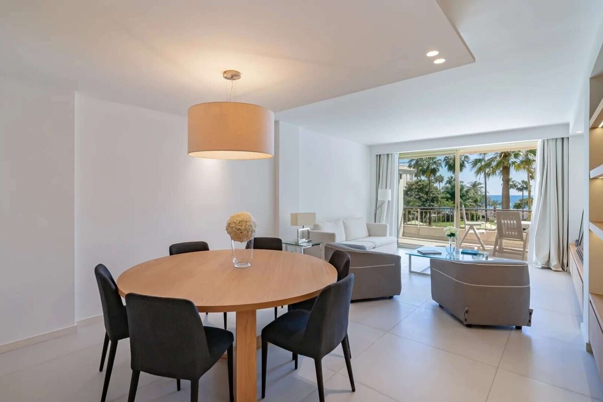 Bright, modern dining and living room with a round table, sofa, and glass coffee table, opening to a balcony with palm trees and sea view.